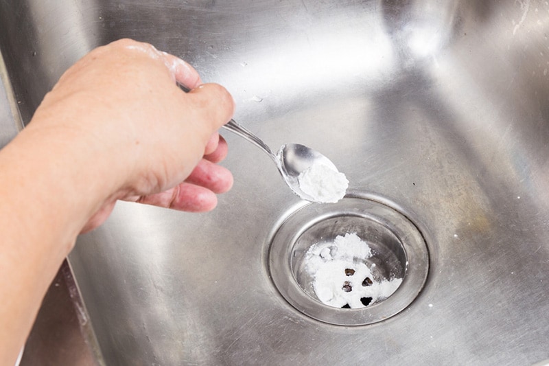 hand holding spoon pouring powder down a kitchen sink drain.