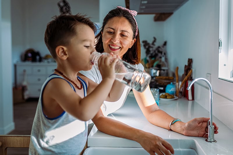 A young boy stands in a kitchen, tilting back a clear glass bottle to drink water. Behind him, a smiling woman with a pink-and-white patterned headband leans over the white countertop, watching him affectionately.