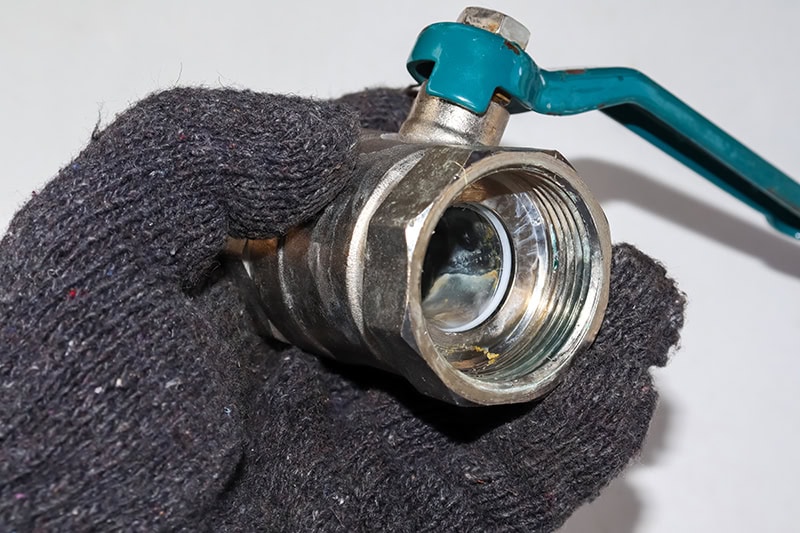 A gloved hand is holding a metal ball valve with a teal-green handle against a plain, light-colored background.