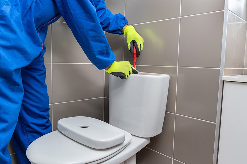 A technician in blue coveralls and yellow gloves repairing a toilet flush mechanism with a screwdriver in a tiled bathroom.