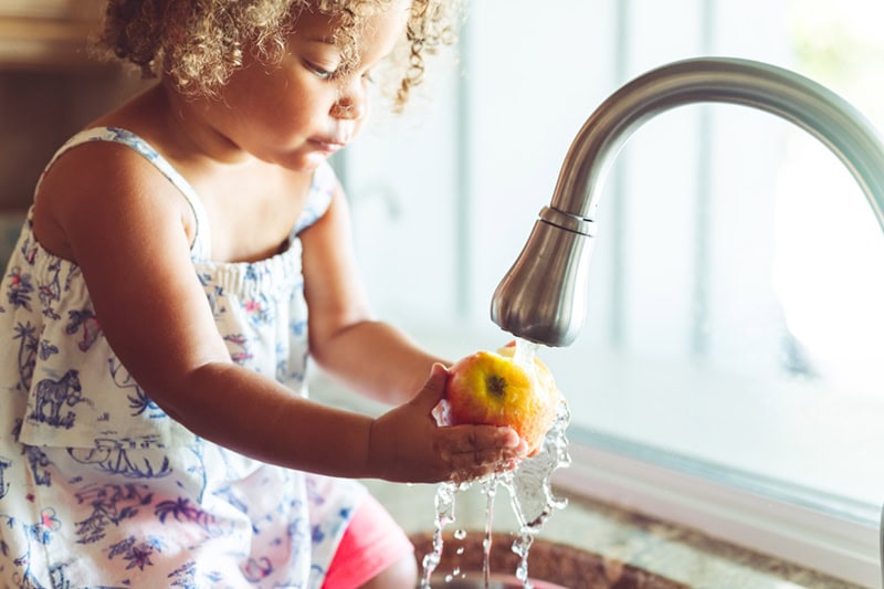 A young child with curly hair is sitting or standing by a kitchen sink, carefully washing a red and yellow apple under running water from a curved, brushed-metal faucet.